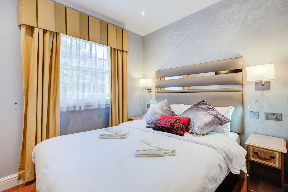 Een hotelkamer met een groot bed, twee nachtkastjes, een raam met gordijnen en een wandlamp.