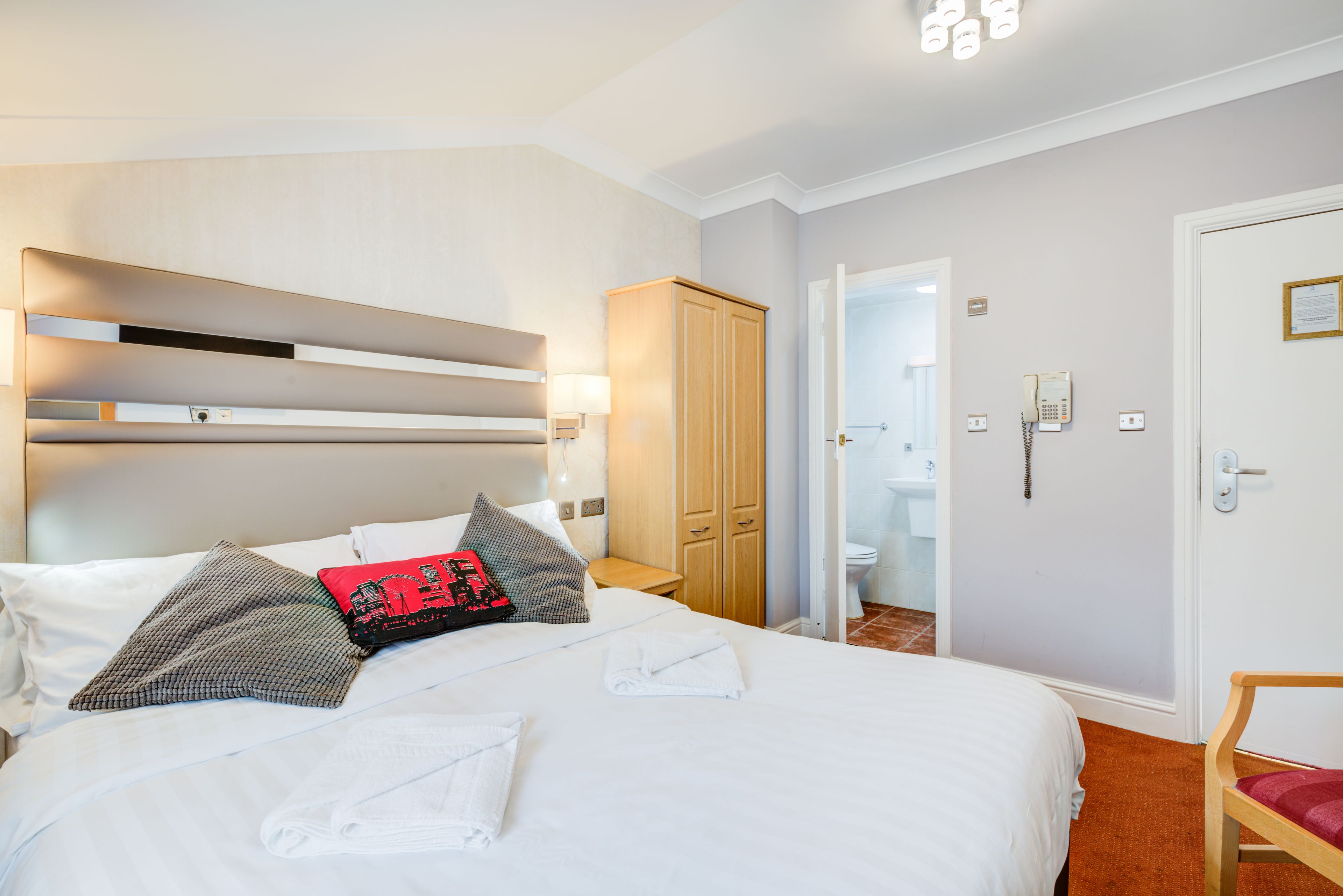 A neatly made bed with white linens and a few pillows, a wooden wardrobe, a bathroom with a toilet and sink, and a chair with a red cushion.