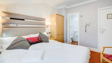 A neatly made bed with white linens and a few pillows, a wooden wardrobe, a bathroom with a toilet and sink, and a chair with a red cushion.