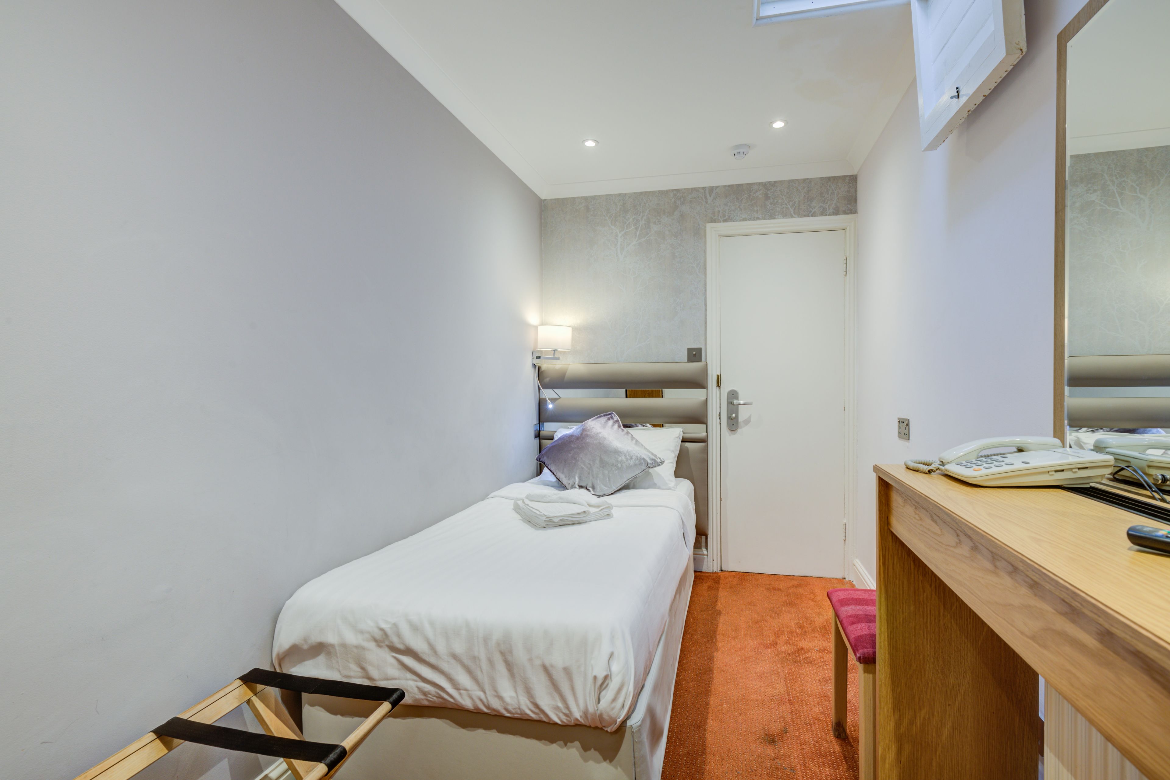A small, neatly arranged bedroom with a single bed, a bedside table with a lamp, and a wooden desk with a telephone.