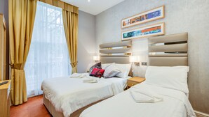 Een hotelkamer met twee bedden, een bureau en een raam met gordijnen. Er hangen afbeeldingen in lijsten aan de muur en een lichtschakelaar.