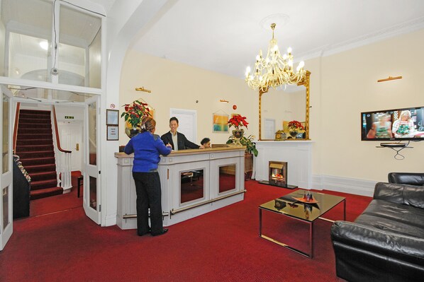 A reception area with a counter, a chandelier, a television, and a staircase. - Admiral Hotel at Park Avenue (London)
