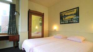 Habitación de hotel con dos camas, un escritorio de madera, un espejo y un cartel en la pared del Hotel Lugano Grand & Palace.