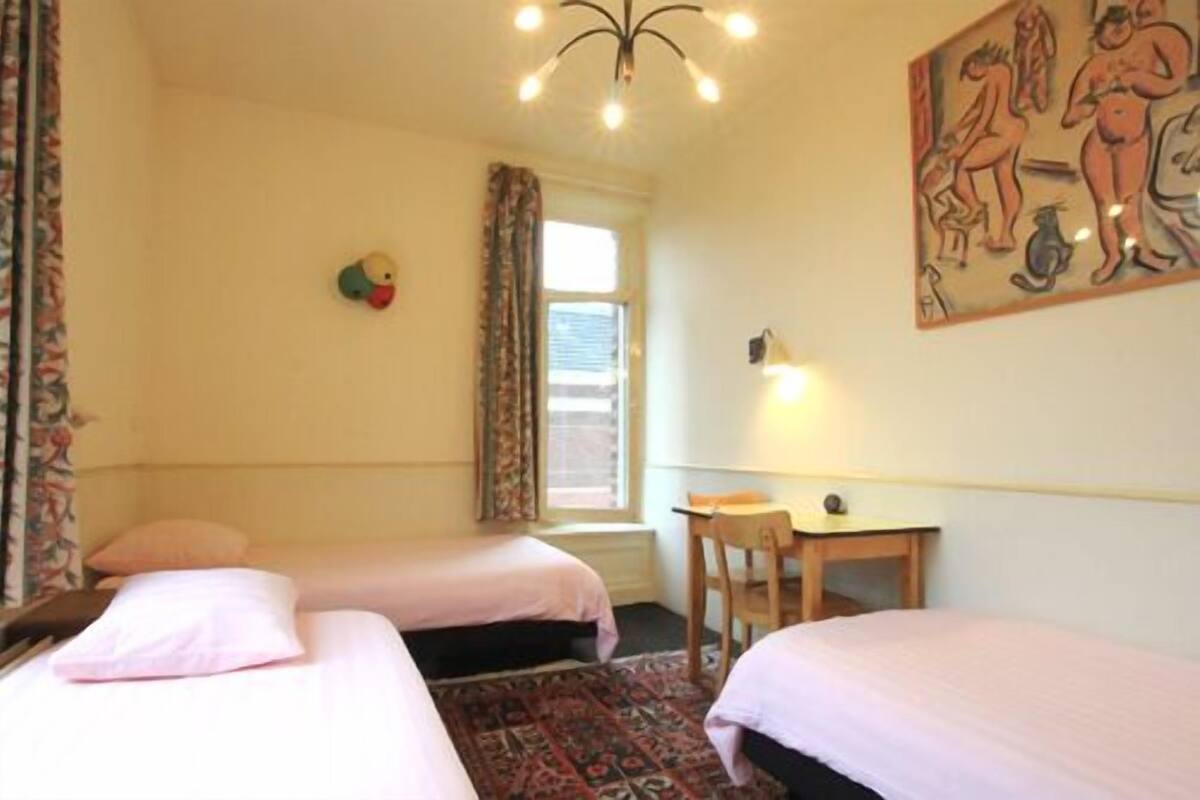 A room with two beds, a small table, a chair, a window with curtains, and a painting on the wall.
