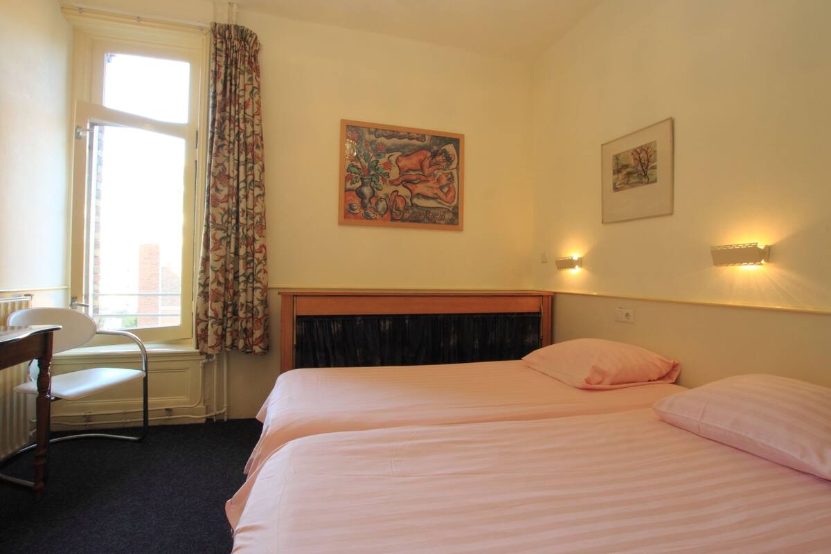 A hotel room with two beds, a desk, a chair, a window with curtains, and wall-mounted lights.