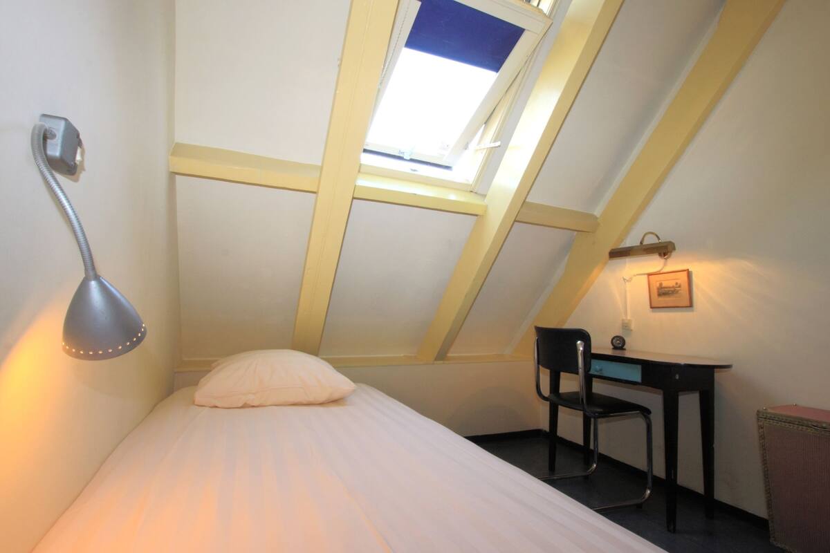 A small, single-bed room with a sloped ceiling, a desk, a chair, and a wall-mounted lamp.