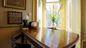 Una mesa de madera con un cuenco y un objeto decorativo, una obra de arte enmarcada en la pared, y una ventana con vistas a la calle y al verdor.