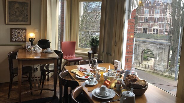 Un área de comedor con una mesa dispuesta para el desayuno, una silla roja y vistas a un edificio histórico a través de amplios ventanales.