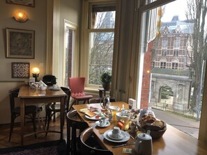 A dining area with a table set for breakfast, a red chair, and a view of a historic building through large windows. - Hotel Museumzicht (Amsterdam)