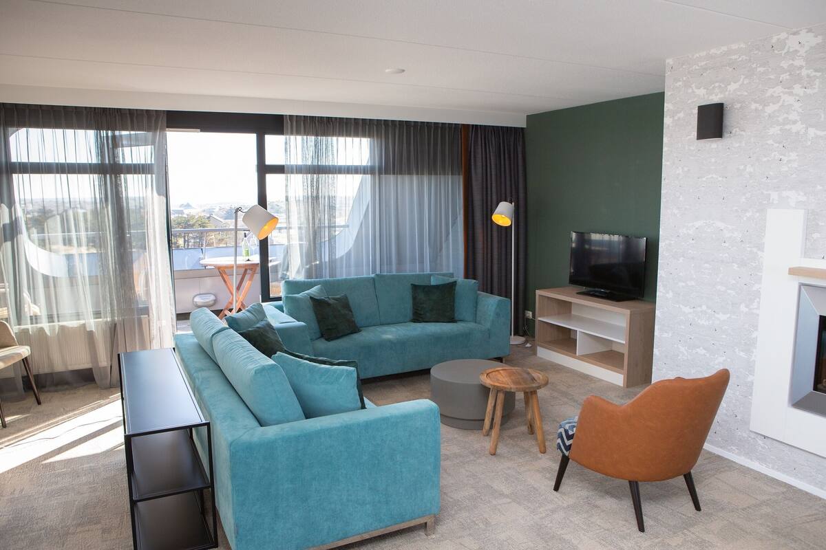 Ein modernes Wohnzimmer mit Sofa, Couchtisch und Sesseln, einem Fernseher und Blick durch große Fenster.