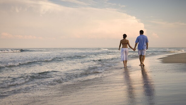 Ein Paar spaziert am Strand, im Hintergrund sind Wellen zu sehen.