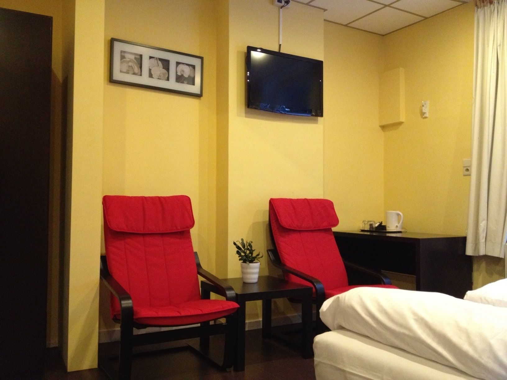 Habitación de hotel con dos sillas rojas, una pequeña mesa con una planta, un televisor en la pared y una cama con ropa de cama blanca.