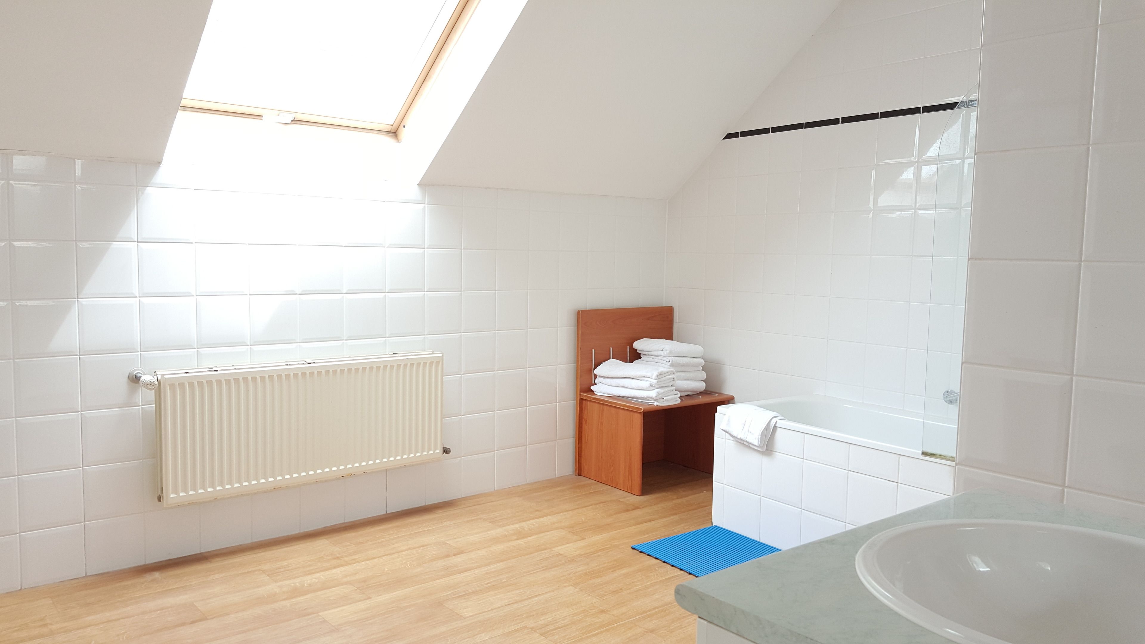 Un baño moderno con baldosas blancas, suelo de madera, tragaluz, radiador, estante de madera con toallas y bañera.