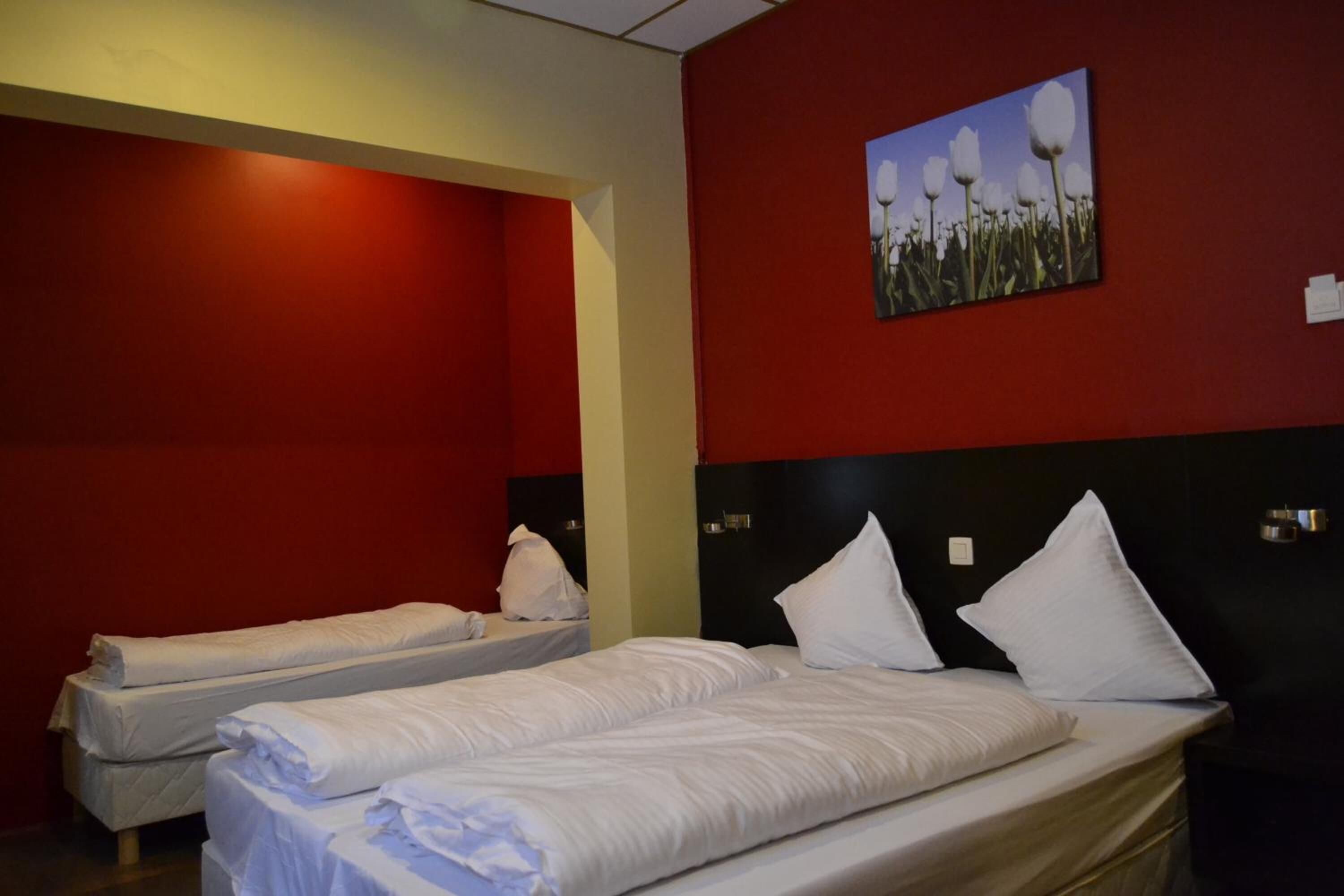 A hotel room with two single beds, a red accent wall, and a painting of flowers.