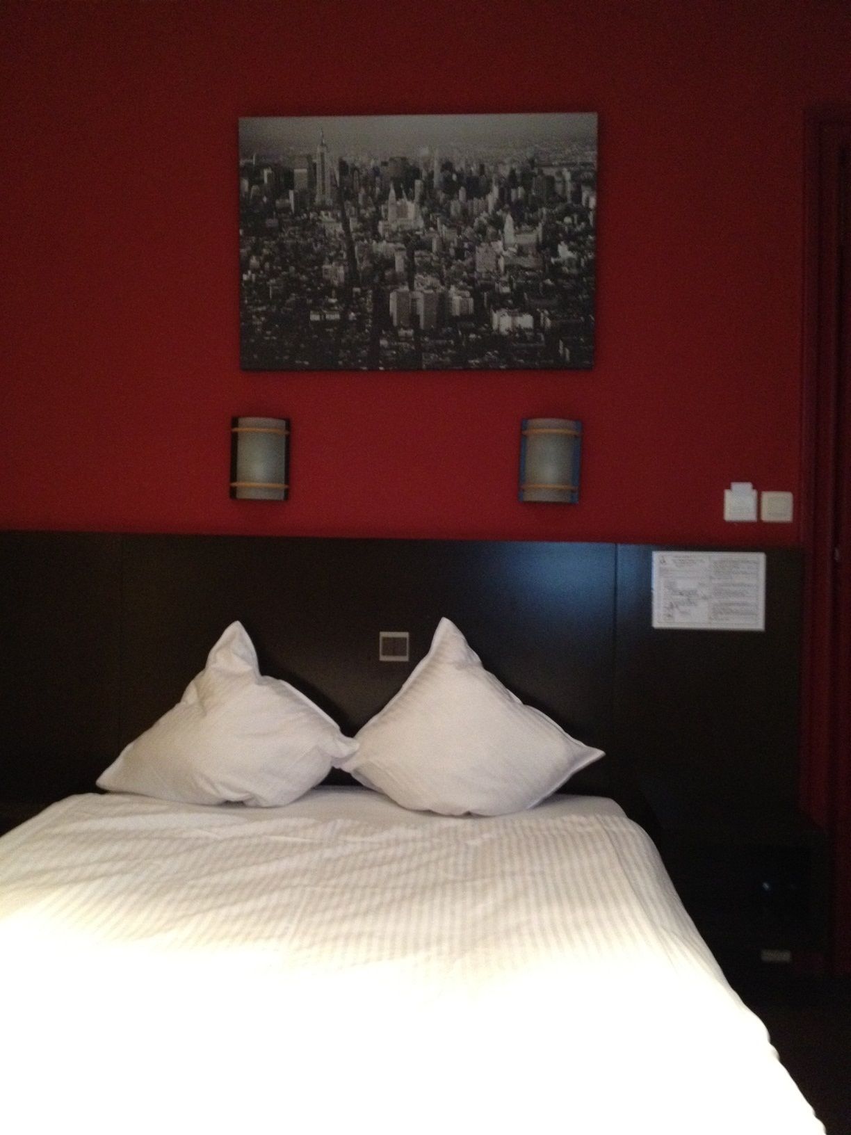 A hotel room with a large bed, two pillows, a headboard, a cityscape picture on the wall, two wall sconces, and a small sign on the wall.