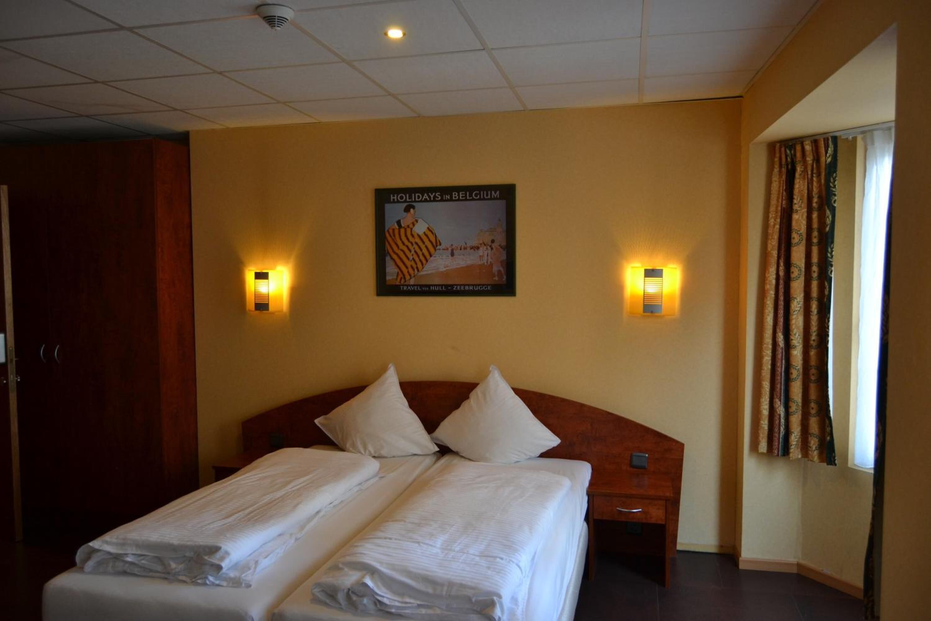 A hotel room with a double bed, two bedside tables, a wooden headboard, two wall-mounted lamps, and a poster on the wall advertising holidays in Belgium.