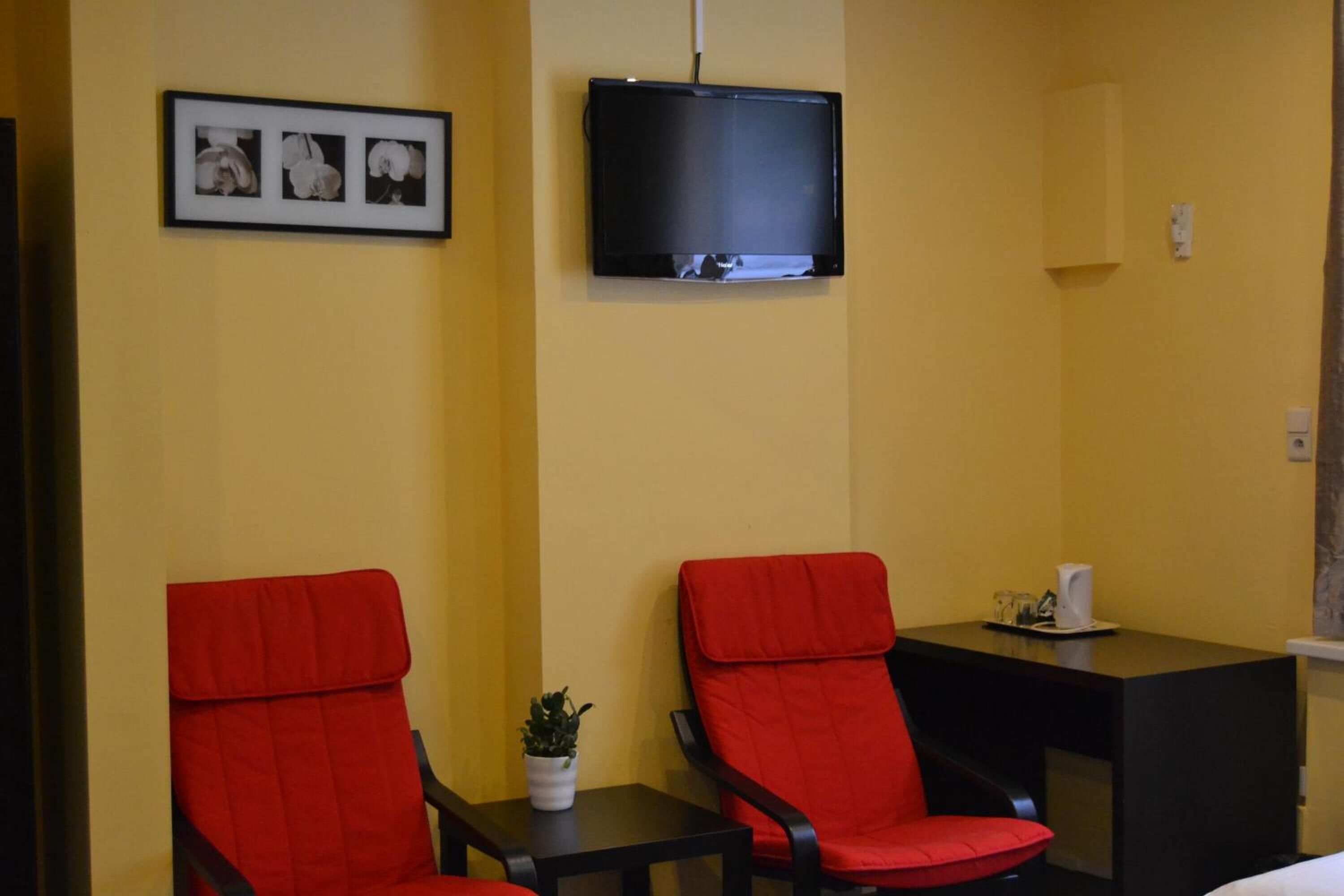 A hotel room with two red chairs, a small table with a plant, a flat-screen TV mounted on the wall, and a framed picture above the TV.