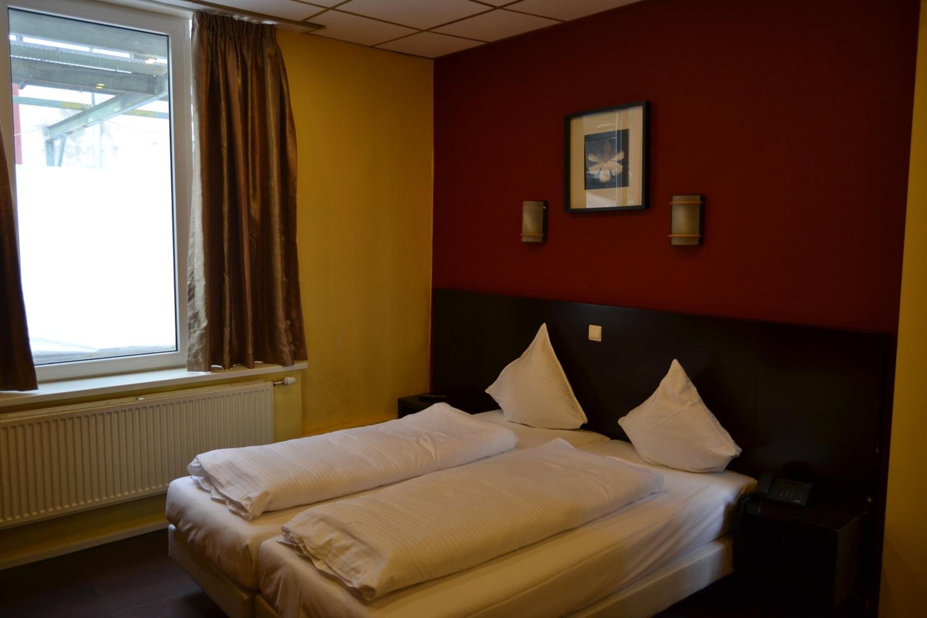 Een hotelkamer met een bed, een raam met gordijnen, een radiator en een aan de muur hangend schilderij.