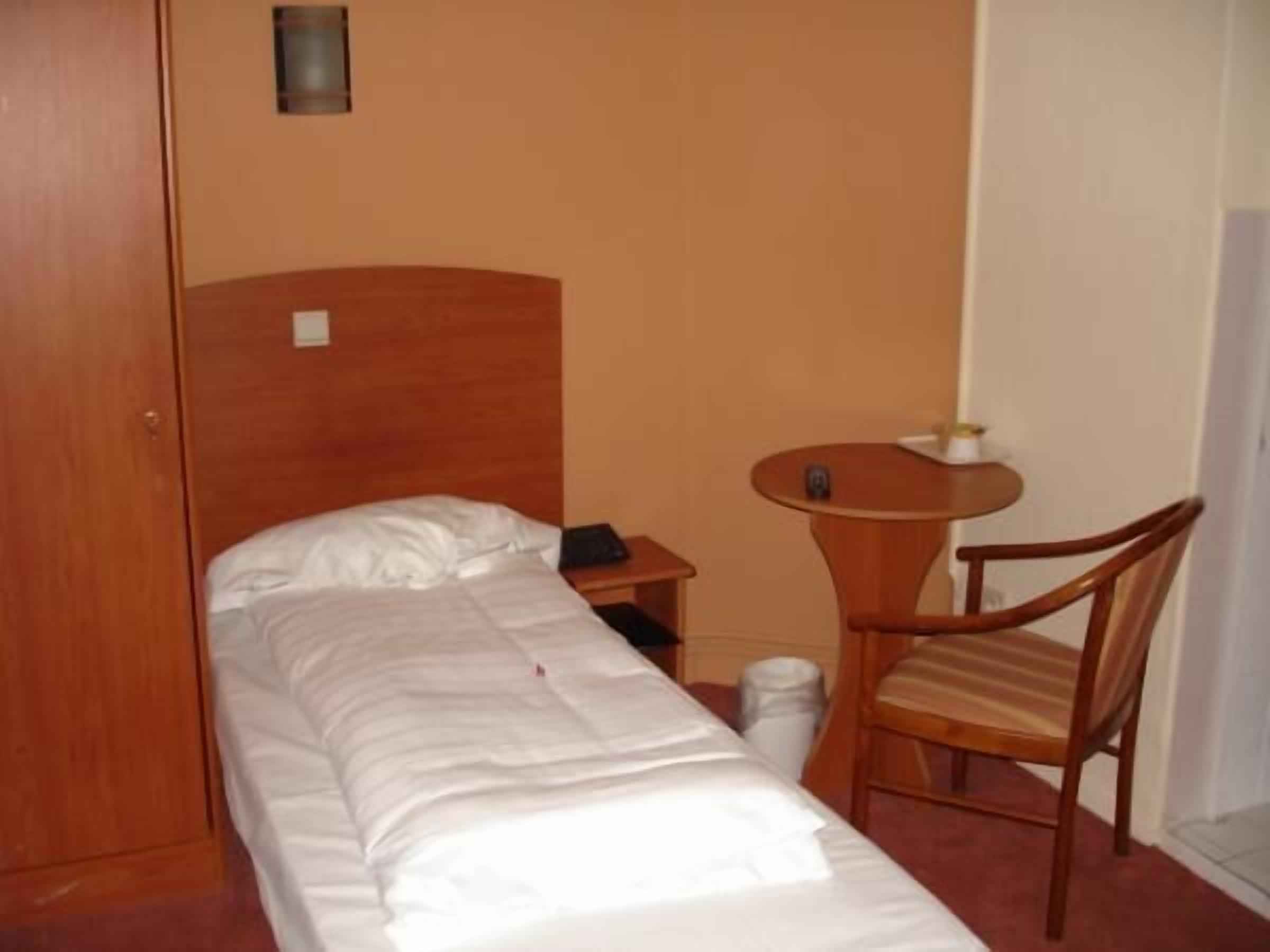Una cama individual con cabecera de madera, una mesita de noche con lámpara y una silla.