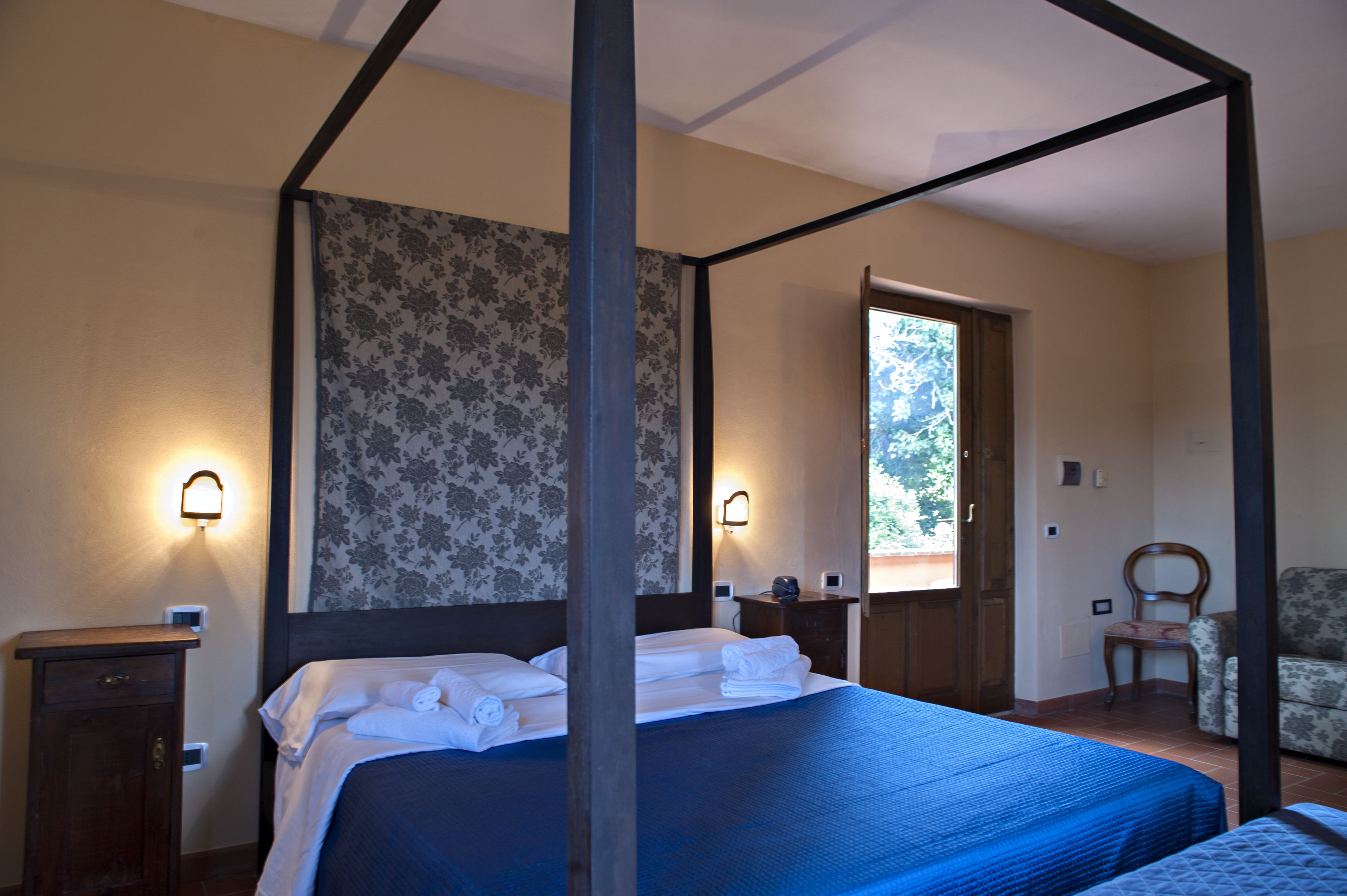 Ein Bett mit Baldachin, mit blauer Bettdecke, ein Nachttisch aus Holz, ein Stuhl und eine Tür, die auf einen Balkon führt.