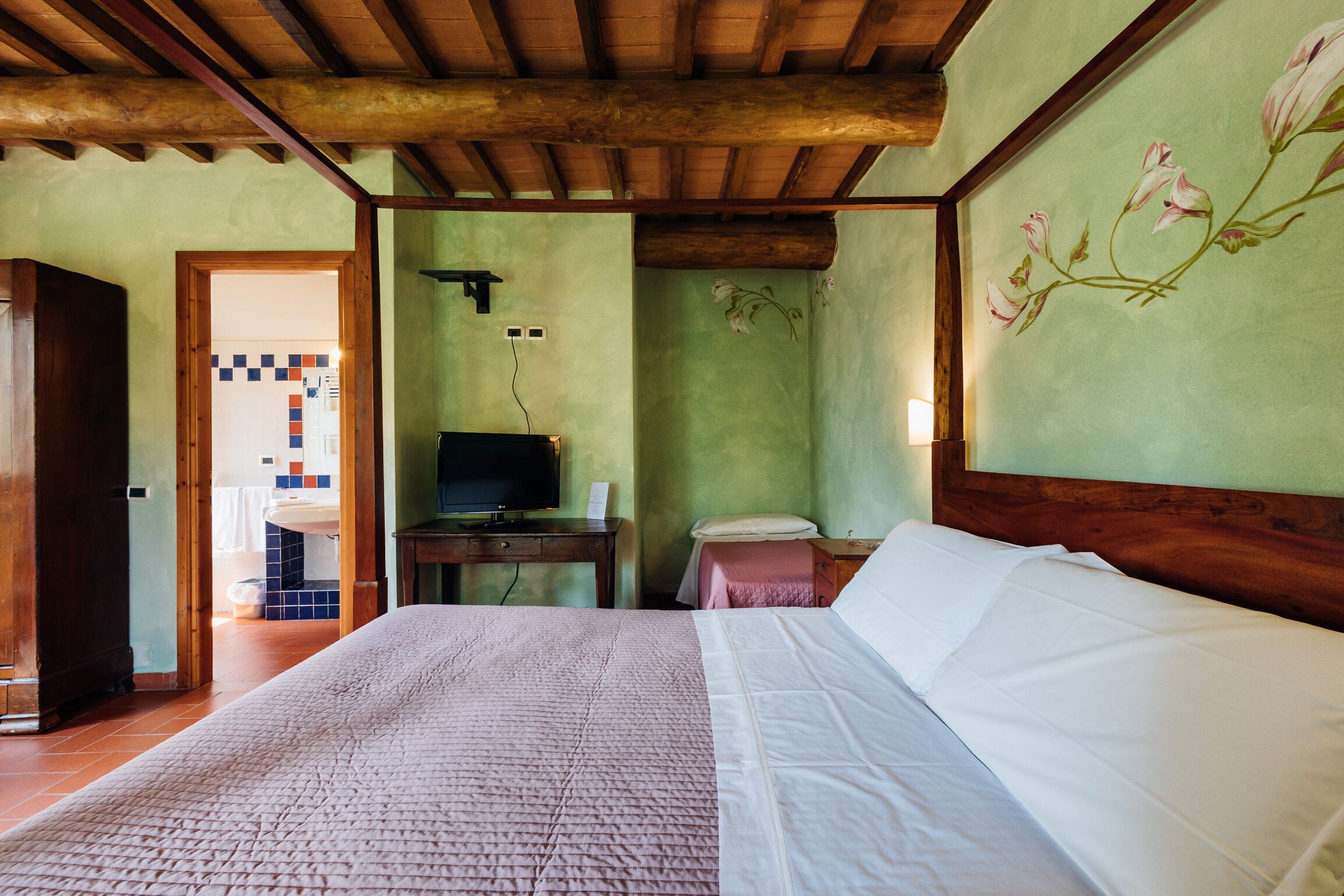 Un dormitorio con techo de madera, una cama con ropa de cama blanca, un televisor sobre una mesa de madera, y un baño visible a través de una puerta abierta.