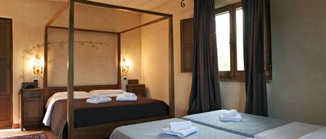 A four-poster bed with a wooden frame and a dark-colored bedspread. There are two bedside tables with lamps and a window with curtains.