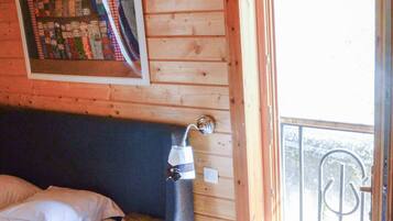 A wooden cabin room with a bed, a table with a book, a lamp, and a window with a view of greenery.