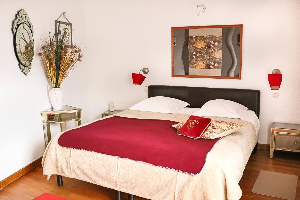 Une chambre à coucher bien rangée, avec un lit, une table de chevet, une commode, un vase contenant des plantes séchées et deux lampes fixées au mur.