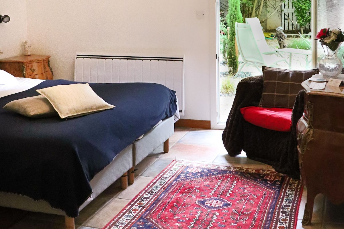 Une chambre avec un lit, un radiateur, une fenêtre donnant sur un espace verdoyant, une chaise et une table sur laquelle se trouve un vase.