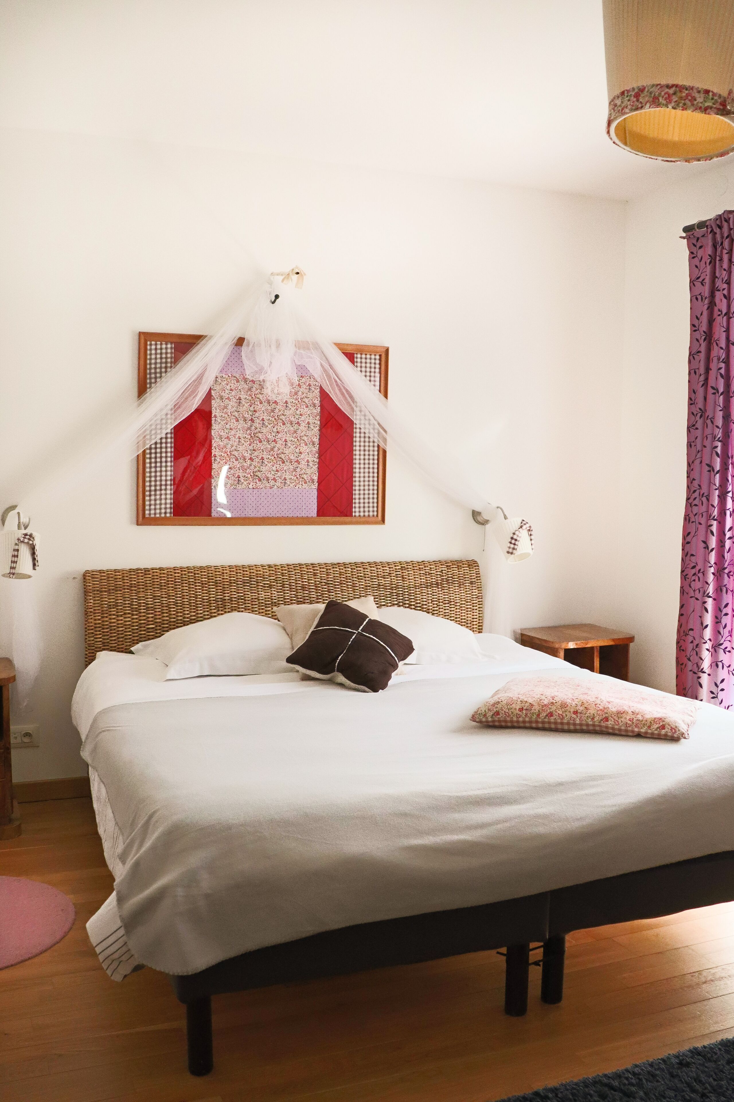 Quarto com cama, mesa de cabeceira, pequena mesa, luminária na parede e um objeto decorativo sobre a cama.