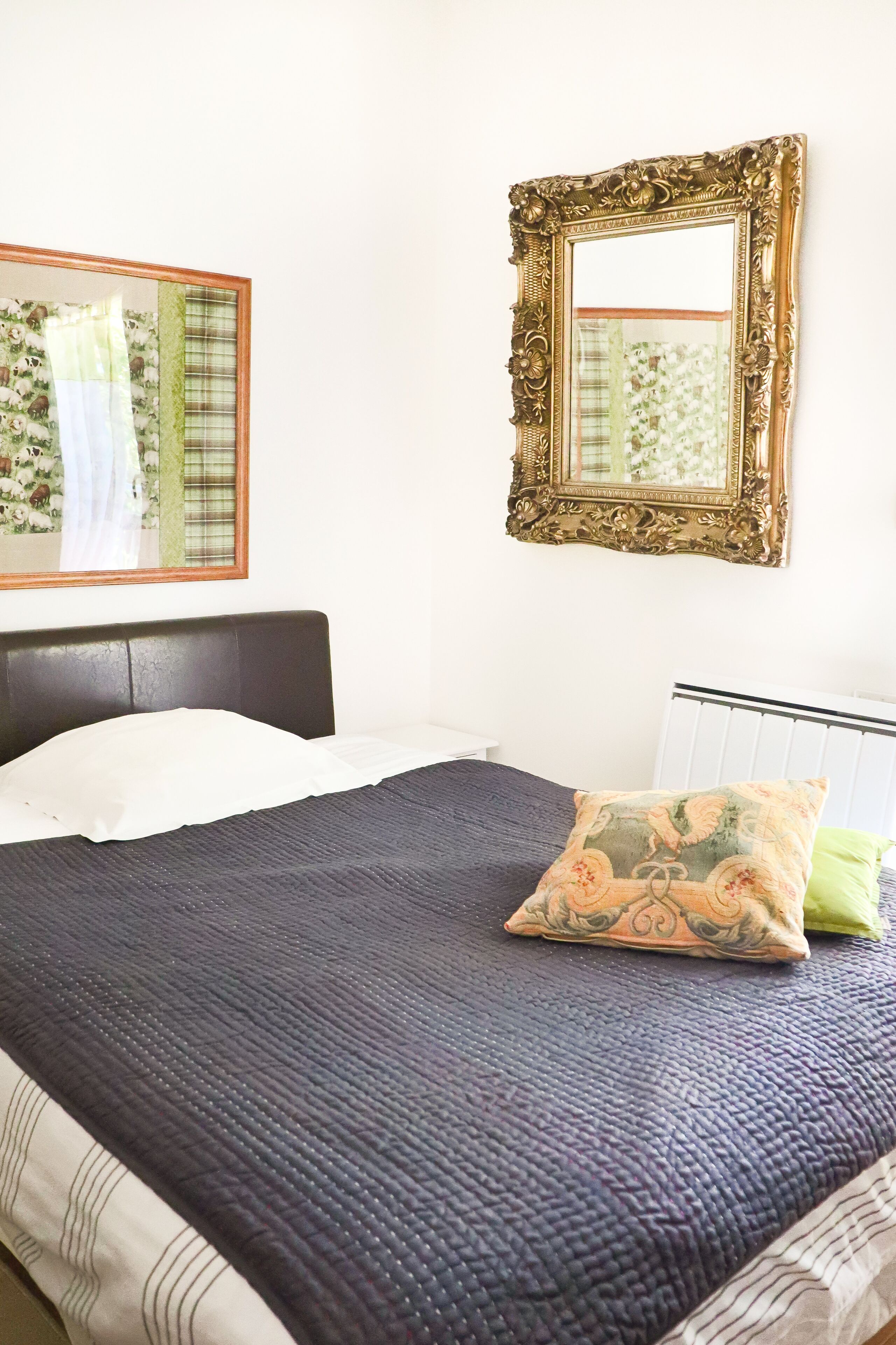 Un lit bien fait, avec une couverture bleu foncé et un coussin décoratif, une tête de lit blanche et un miroir mural doté d’un cadre orné.