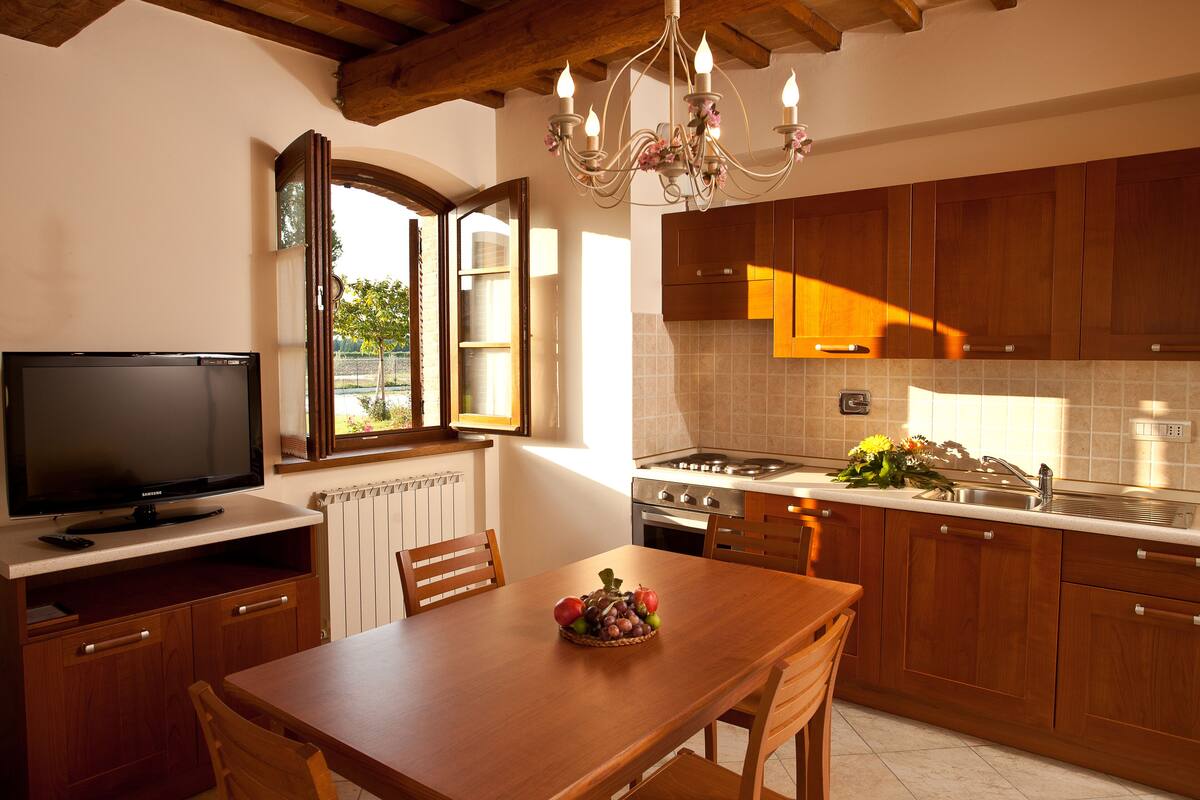 A kitchen with wooden cabinets, a dining table with chairs, a chandelier, and a television on a stand.