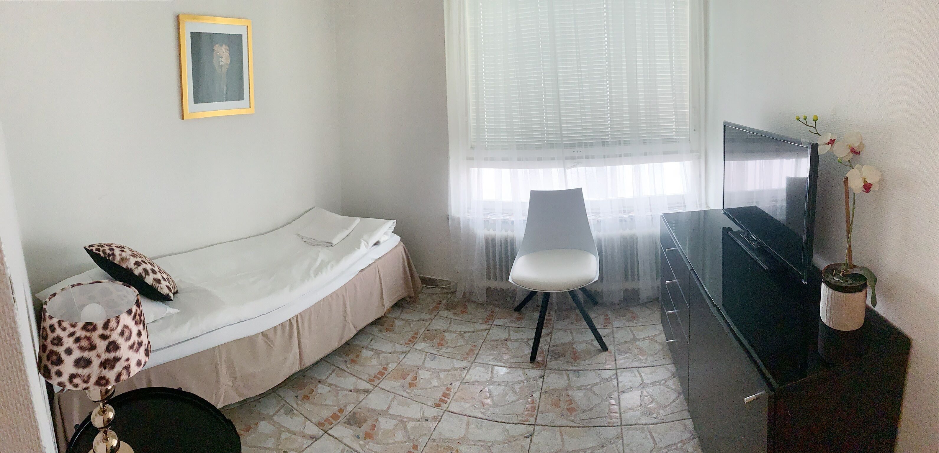 Une petite pièce comprenant un lit, une chaise, un téléviseur et une fenêtre avec des stores.