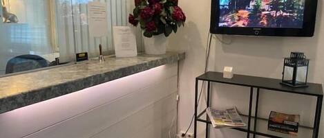 Una moderna zona de recepción de hotel con un mostrador de mármol, un televisor montado en la pared, un jarrón con flores rojas y una pequeña mesa negra con revistas.