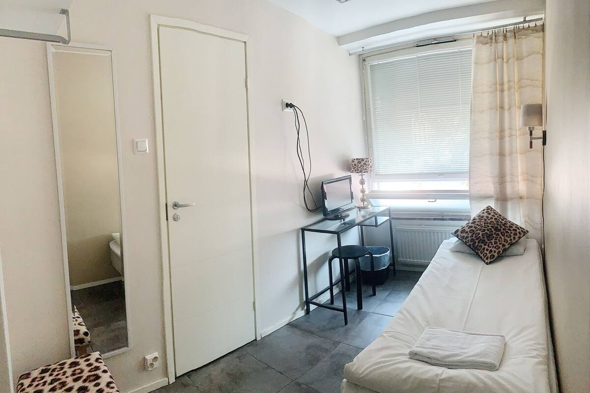 A room with a bed, a desk with a computer, a chair, a radiator, and a window with curtains.