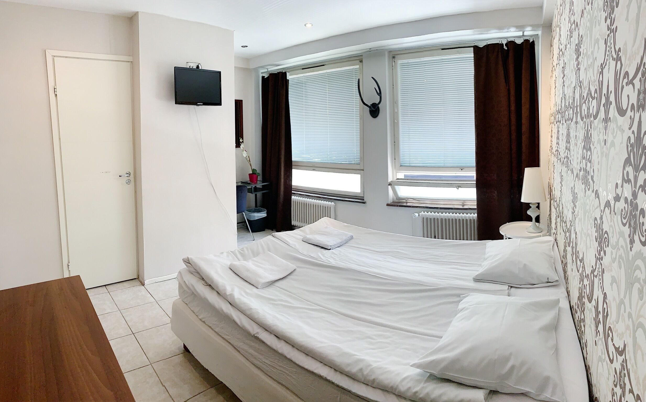 Hotellihuone, jossa on sänky, seinälle asennettu televisio, patteri ja ikkuna, jossa on verhot.