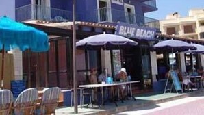 A hotel with outdoor seating and umbrellas, a sign reading "Blue Beach," and a clear sky. - Blue Beach (CAN PICAFORT)