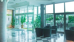 Eine großzügige Lobby mit Schachbrettmuster-Boden, großen Fenstern mit Blick nach außen und mehreren Blumentöpfen.