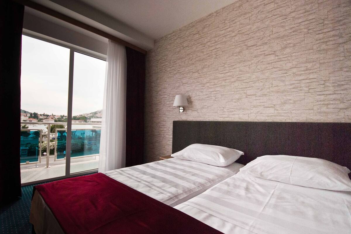Hotellihuone, jossa on suuri sänky, yöpöytä, lamppu ja näkymä uima-altaalle ja ympäröiville rakennuksille.