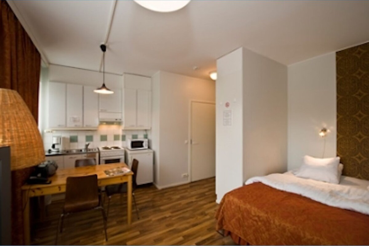 A compact hotel room with a wooden floor, a bed with white bedding, a small dining table with chairs, a kitchenette, and a hanging lamp.