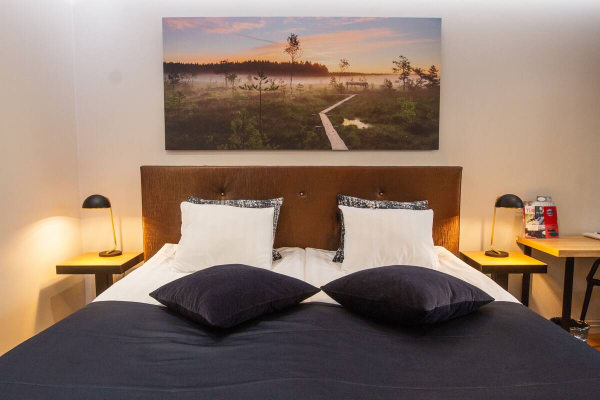 Una cama bien tendida con ropa de cama blanca y azul oscuro, dos lámparas de noche y un cuadro de un paisaje en la pared.