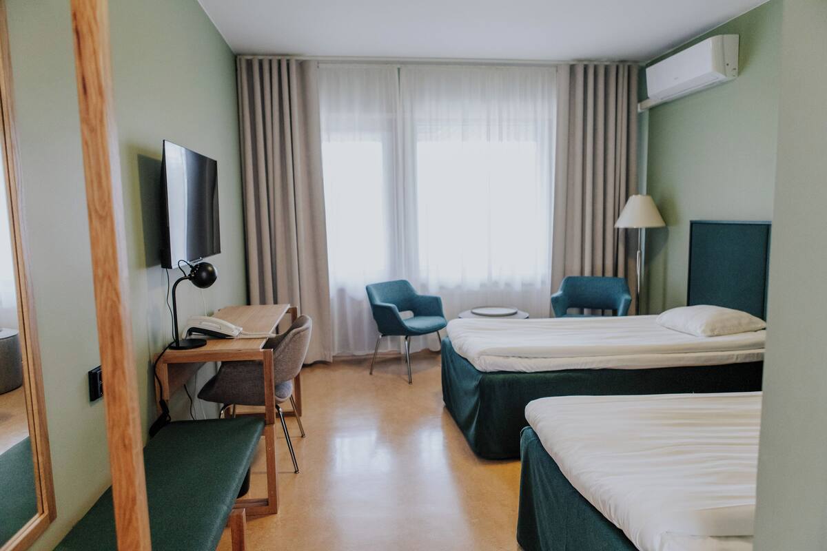 Una habitación de hotel con dos camas, un escritorio con silla, televisión, lámpara y ventana con cortinas.