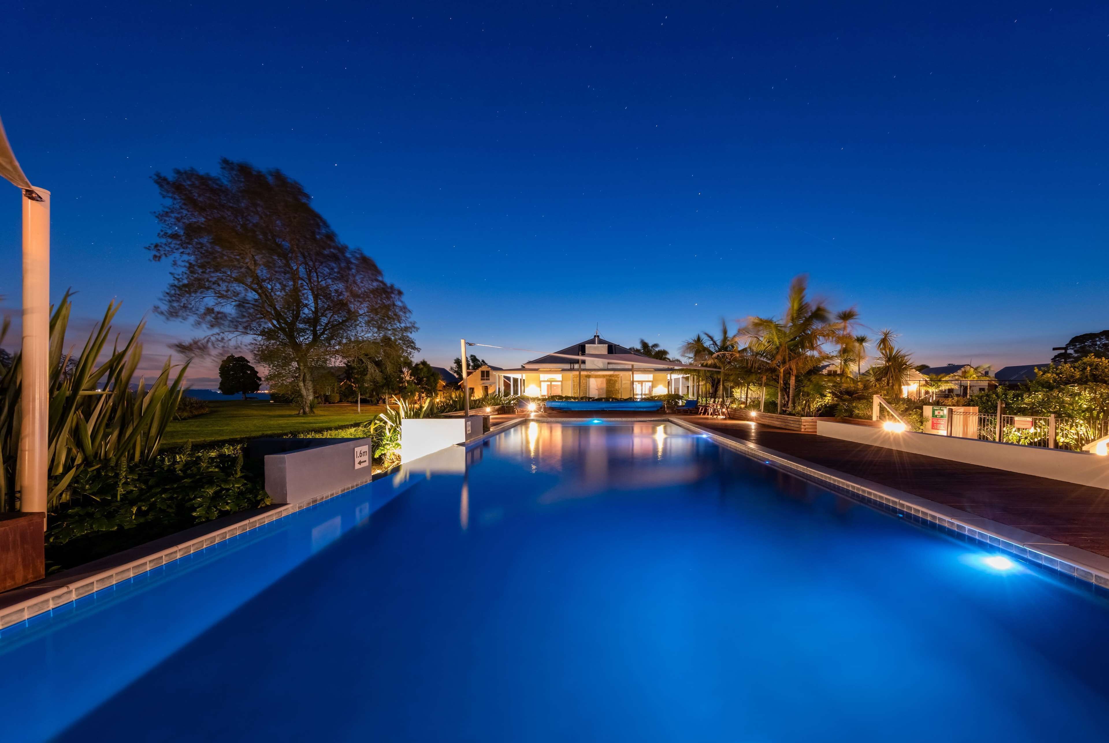 Vue nocturne d’un espace piscine avec une maison en arrière-plan, entourée de palmiers et de chemins bien éclairés.