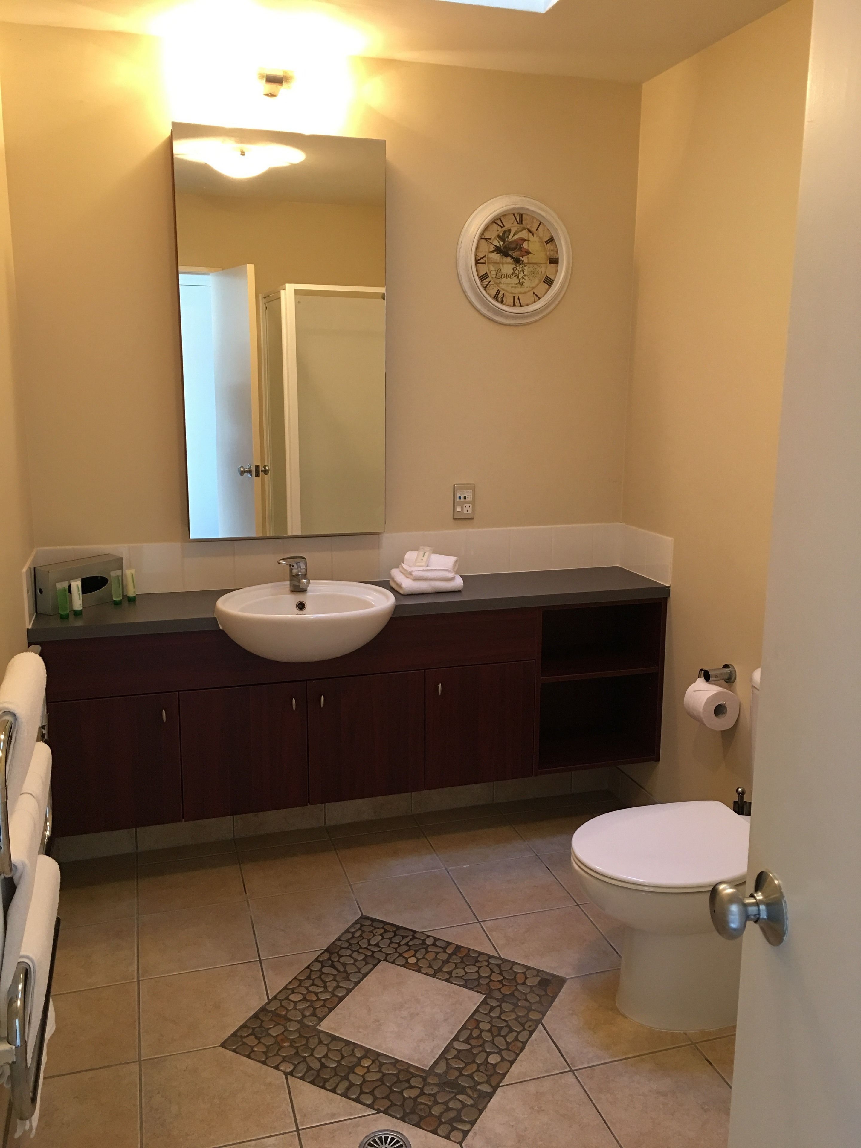 A bathroom with a white sink, dark wooden cabinets, a round mirror, a clock, and a toilet.