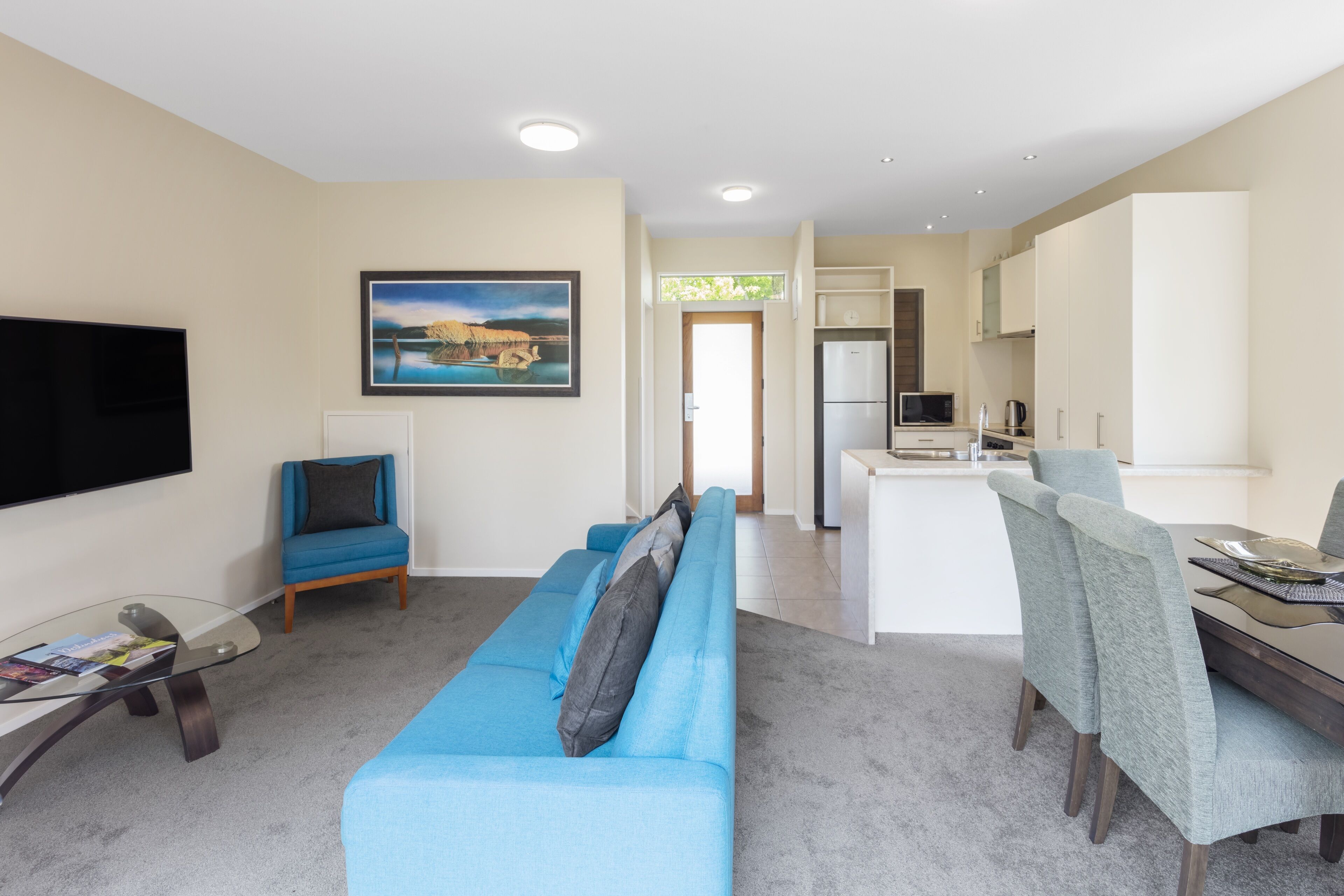 A modern living room with a blue sofa, a glass coffee table, and a flat-screen TV. There is a dining area with a table and chairs, and a kitchen with white cabinets and appliances.