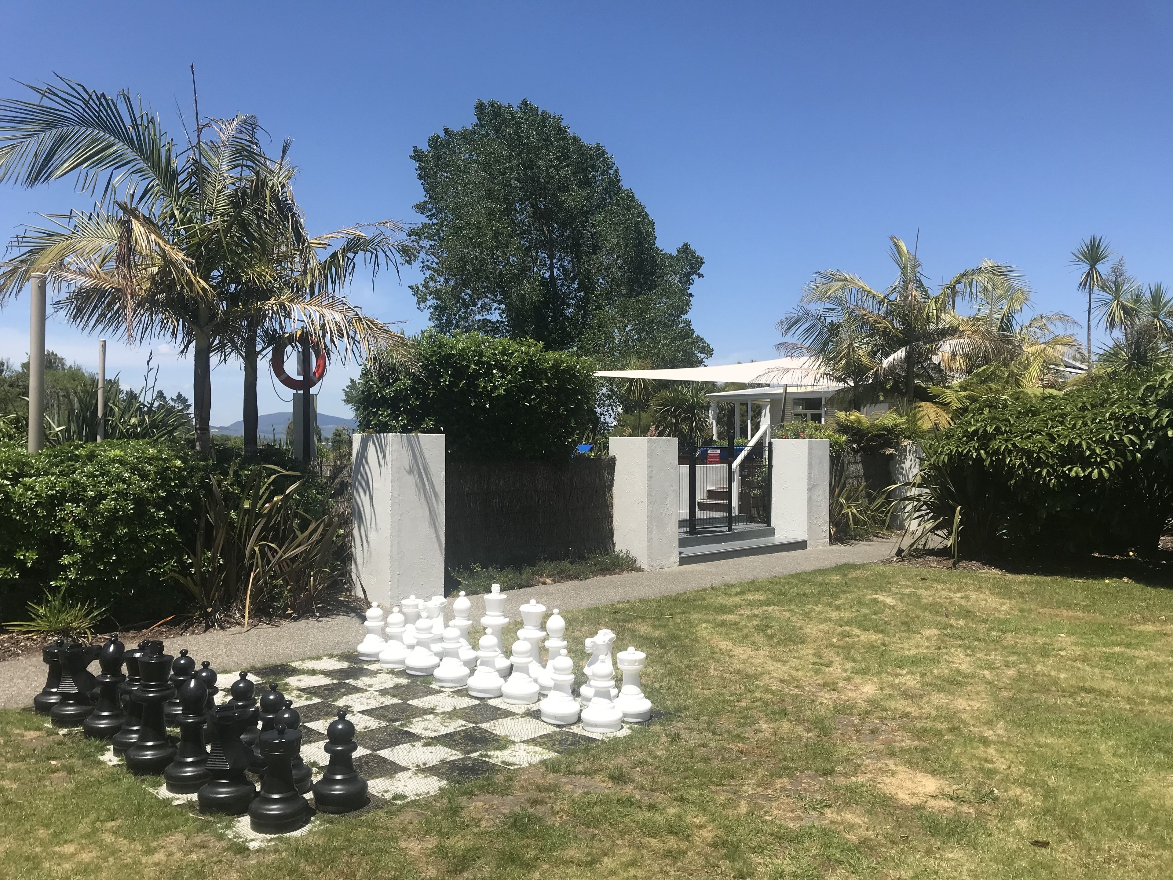 A chessboard set up outdoors with a garden and a building in the background.