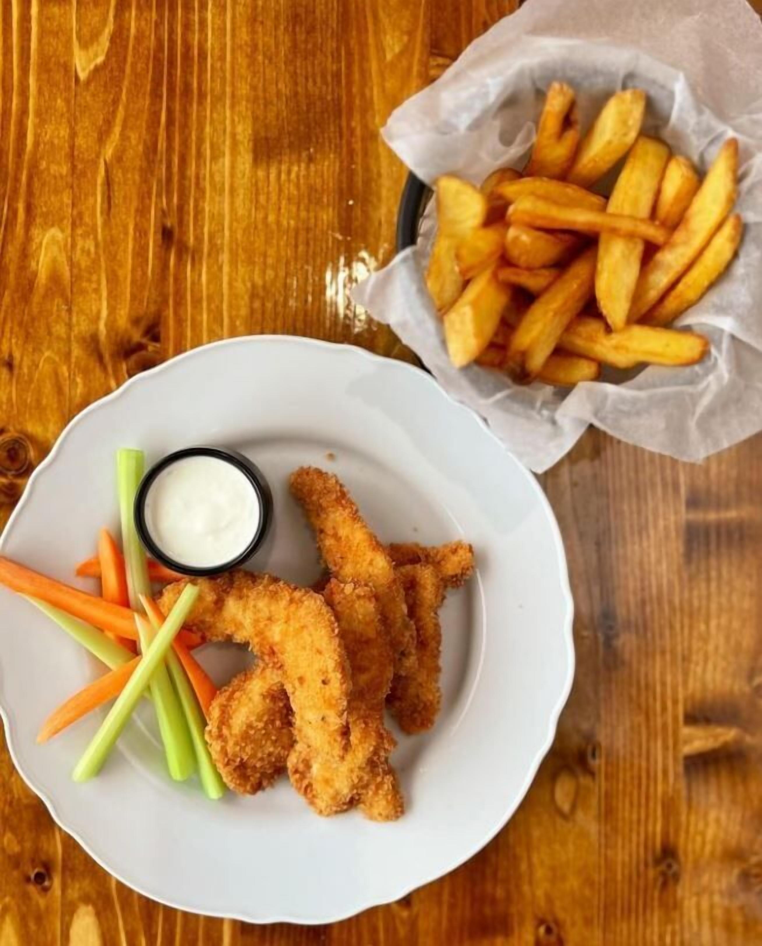 Un plato de tiras de pollo empanizadas y fritas, palitos de apio y zanahoria, acompañados de una salsa para mojar en un recipiente pequeño y una cesta de papas fritas.