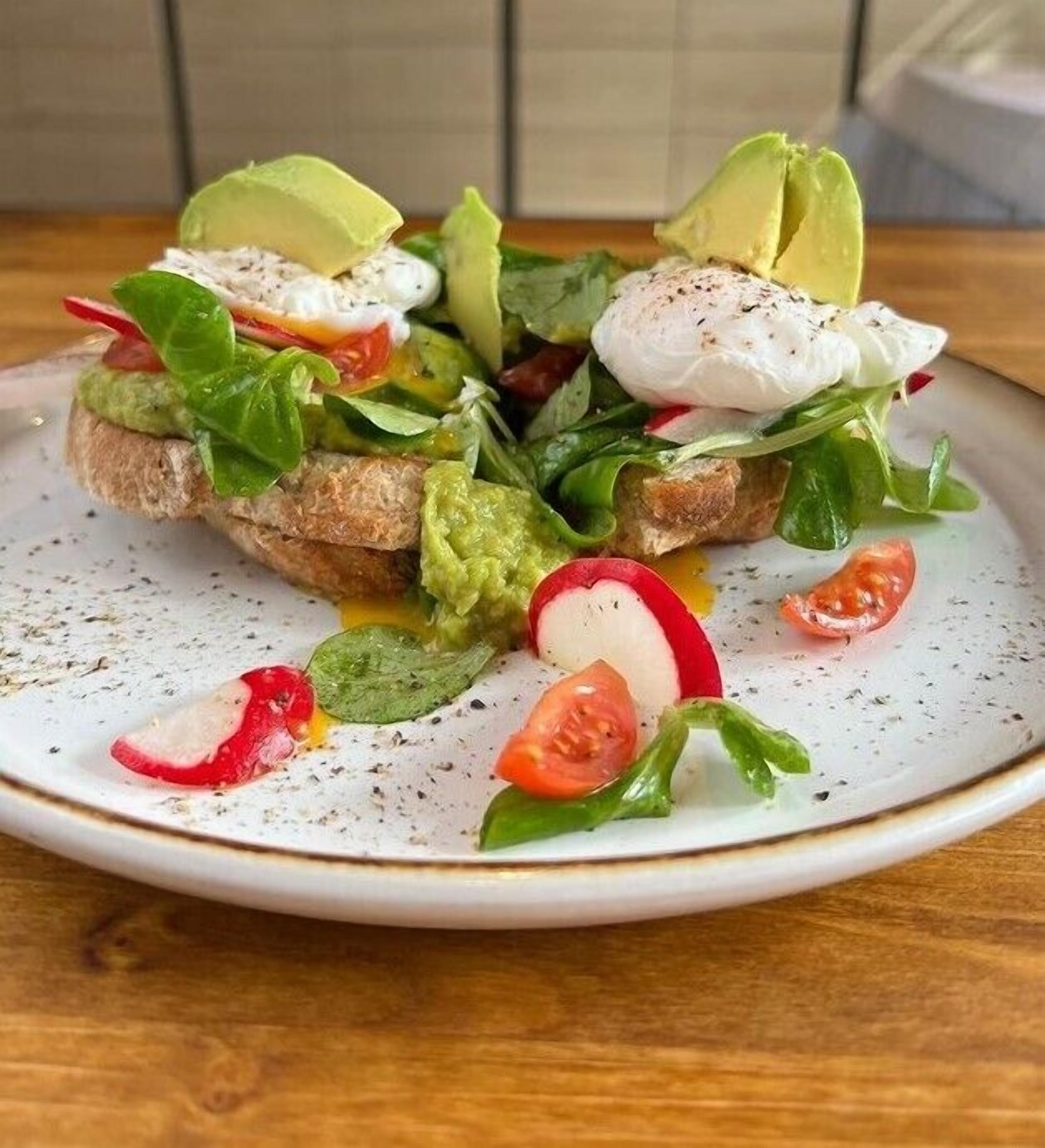 A plate with a sandwich, avocado slices, radish, and tomato on a speckled ceramic plate.