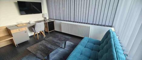 A modern hotel room with a grey sofa, a desk with a chair, a flat-screen TV, and a radiator.