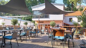 Un espace extérieur aménagé en terrasse, avec des tables et des chaises, ombragé par de grands auvents et entouré d’arbres et de bâtiments.